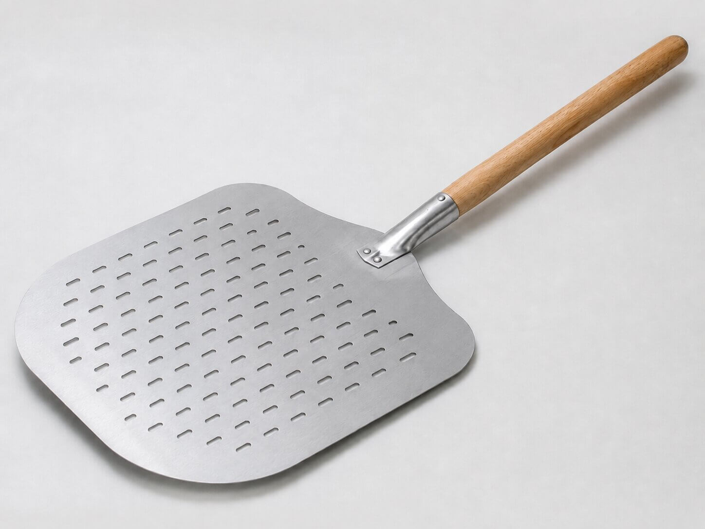 n perforert pizzaspade i aluminium med et rundt treskaft, liggende diagonalt på en lys og nøytral bakgrunn.